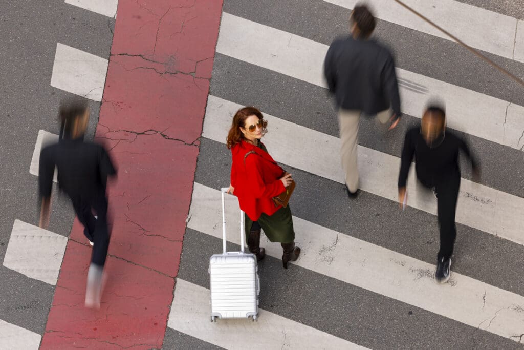 Lady with a red jacket and silver suitcase crossing the road.